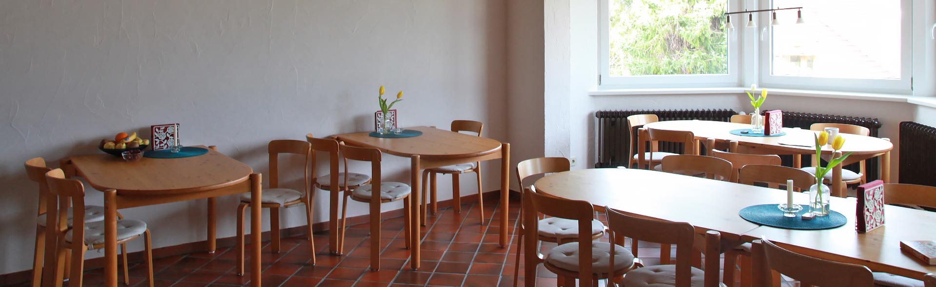 OJC Begegnen – Freundlicher Essraum mit Vollholz-Einrichtung, ziegelroten Bodenfließen, großer Fensterfront und einer zugänglichen Gästeküche.